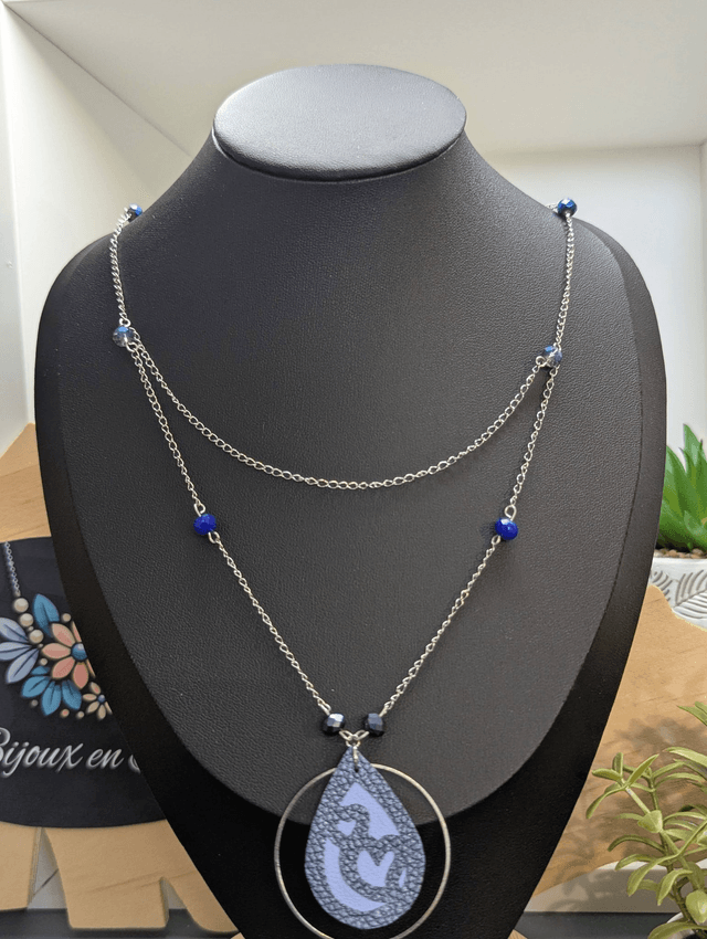 Collier long, sautoir, avec double chaine de couleur argent et un pendentif en cuir et en tissus enduit en forme de goutte et de cœur bleu.