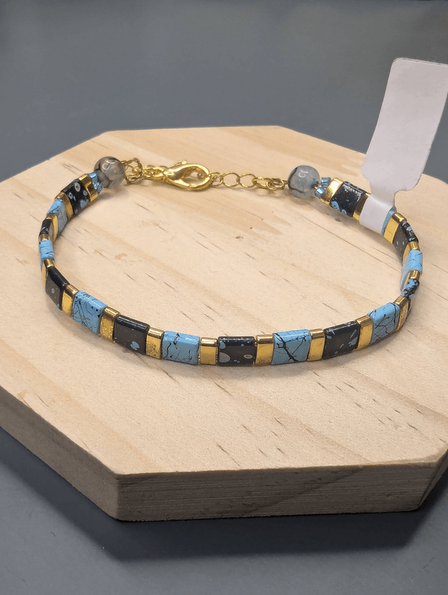 Bracelet avec des perles plate en verre noires et bleus et des perles en métal doré. Le fermoir et la chaine fermoir sont de couleur or.