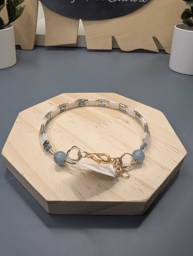Bracelet avec des perles plate en verre blanches et bleu et des perles en métal doré. Le fermoir et la chaine fermoir sont de couleur or.