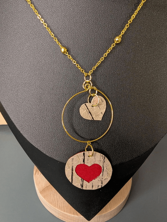 Collier long, sautoir, avec une chaine de couleur or et un pendentif en cuir et en tissus enduit en forme de rond et de cœur.