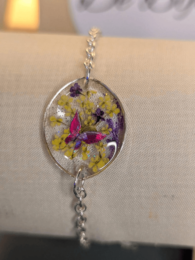 Bracelet avec une chaine de couleur argent, un pendentif en résine avec des fleurs séchés et un papillon en stickers, le fermoir est en argent. 