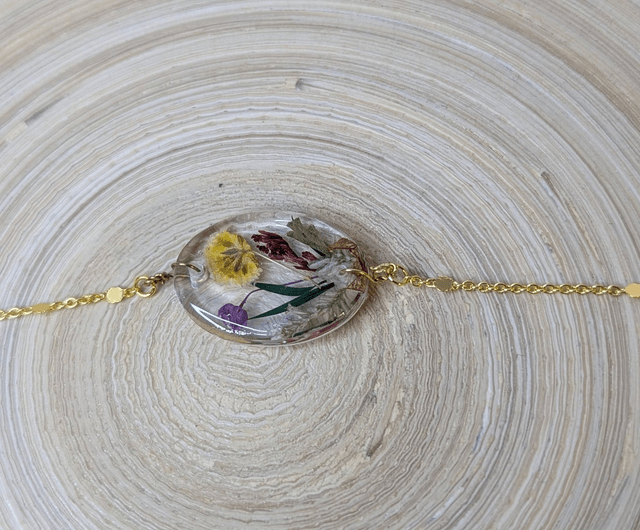 Bracelet avec une chaine de couleur or, le pendentif est en résine avec des fleurs séchés jaune et violet, le fermoir est en or.