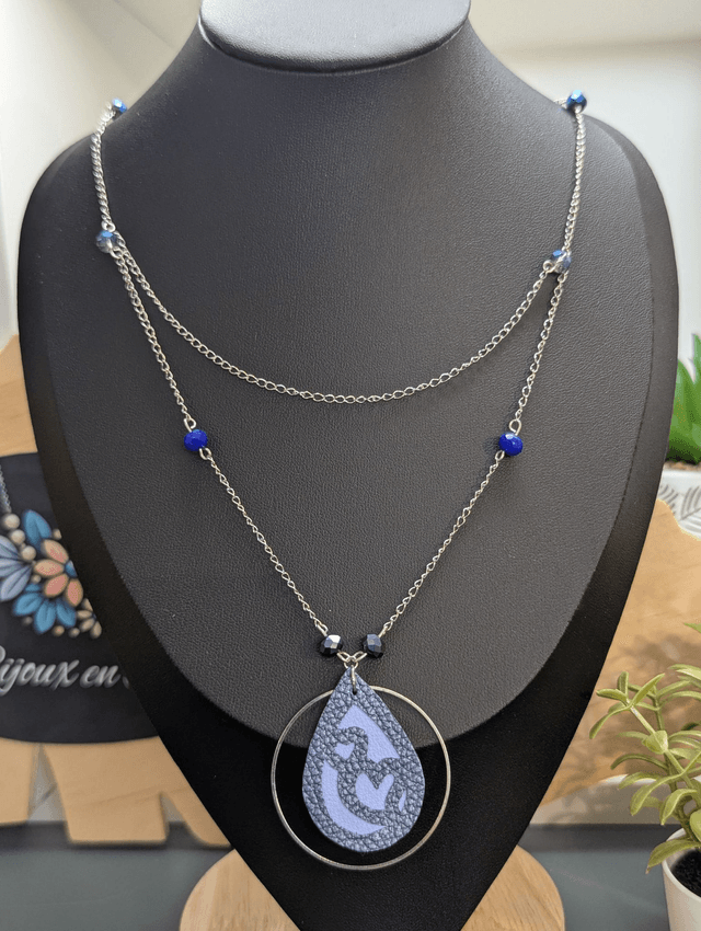 Collier long, sautoir, avec double chaine de couleur argent et un pendentif en cuir et en tissus enduit en forme de goutte et de cœur bleu.