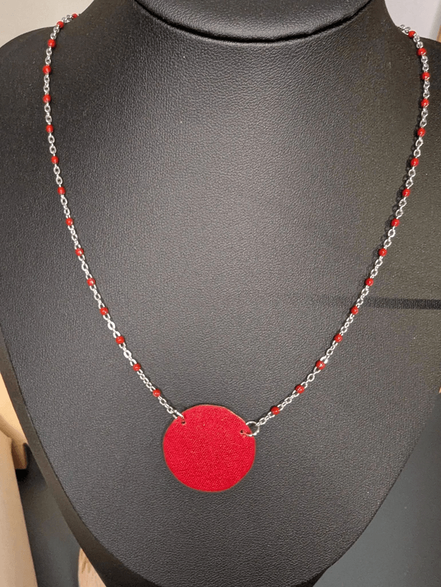 Collier mi long avec une chaine de couleur argent et rouge, avec un pendentif en cuir et en tissus enduit en forme de rond et de cœur. 