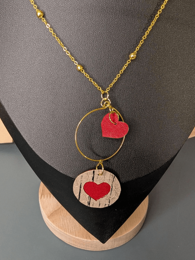 Collier long, sautoir, avec une chaine de couleur or et un pendentif en cuir et en tissus enduit en forme de rond et de cœur.