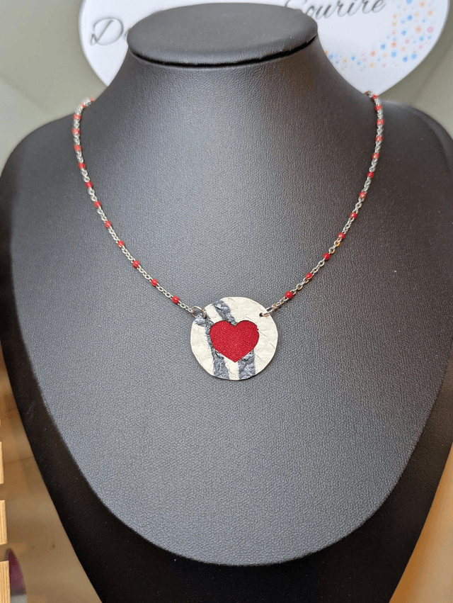 Collier mi long avec une chaine de couleur argent et rouge, avec un pendentif en cuir et en tissus enduit en forme de rond et de cœur. 