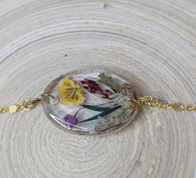 Bracelet avec une chaine de couleur or, le pendentif est en résine avec des fleurs séchés jaune et violet, le fermoir est en or.