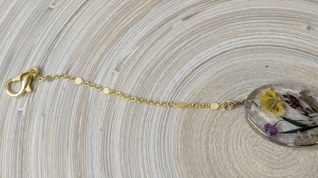 Bracelet avec une chaine de couleur or, le pendentif est en résine avec des fleurs séchés jaune et violet, le fermoir est en or.