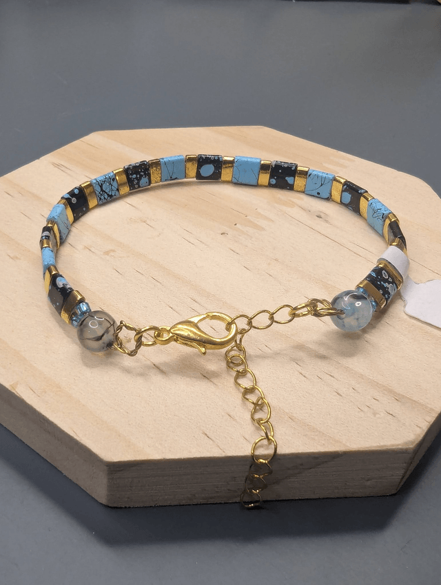 Bracelet avec des perles plate en verre noires et bleus et des perles en métal doré. Le fermoir et la chaine fermoir sont de couleur or.